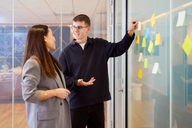 2 colleagues standing in meeting room with glas walls, hanging colourful Post-Its up
